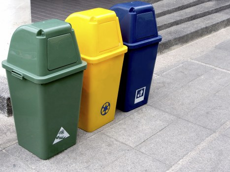 Three upright outdoor rubbish bins positioned on a pavement surface, each made of durable plastic with a smooth, slightly glossy finish. The first bin on the left is forest green with a closed lid, featuring a white recycling symbol and a triangular recycling label on the front; the middle bin is bright yellow with a similarly closed lid, displaying a black circular recycling badge; the third bin on the right is navy blue with a closed lid and a small, white, square label with an icon on the front. They are aligned side by side, close to a building or structure at the edge of the grey, tiled sidewalk. The background includes part of a dark, textured surface, possibly a ramp or step. The environment is well-lit with natural daylight, indicating an outdoor setting in a city area, typical of waste collection points in central locations such as Mayfair, London. The scene reflects professional rubbish disposal and recycling arrangement managed by Business Waste Removal Mayfair, serving commercial and residential premises. The gravelly and smooth finishes of the bins contrast with the textured pavement, emphasizing practical waste management in a busy urban context.
