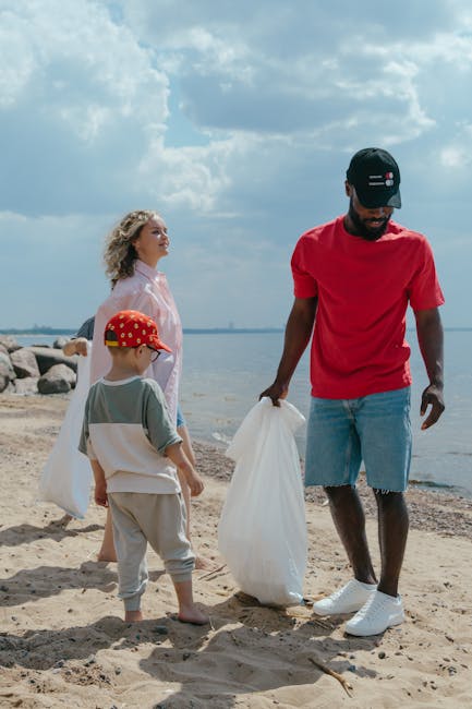 A group of three individuals participating in a beach clean-up activity during daytime, with a man wearing a red t-shirt, light blue denim shorts, and a black cap holding a large white plastic bag. A 