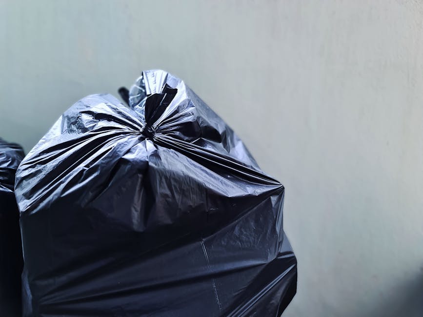 An outdoor urban scene showing multiple overflowing rubbish bins and scattered waste including paper, cardboard boxes, plastic bags, and packaging materials piled around a large grey mixed paper and c